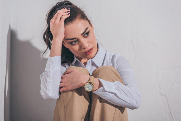 sad woman in blouse and beige pants sitting on white background in room, grieving disorder concept - Photo, Image