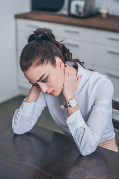 upset woman in white blouse sitting at table in kitchen, thinking and looking down, grieving disorder concept - Photo, Image