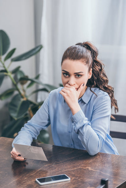 smutná žena v modrá halenka sedí u stolu s smartphone a držení Foto doma, truchlící porucha koncept - Fotografie, Obrázek