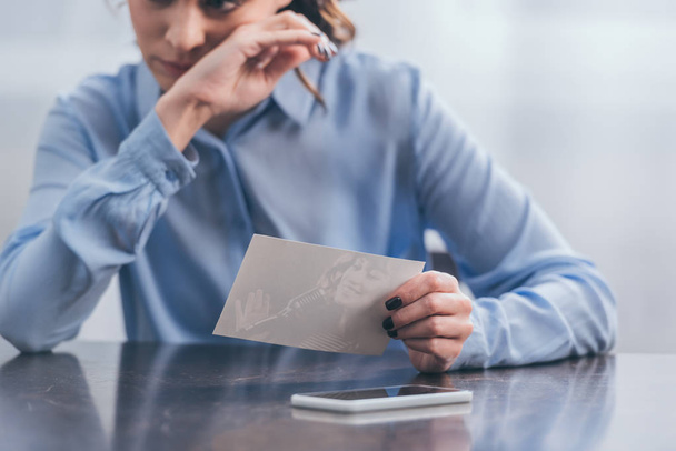oříznutý pohled rozrušená žena v modrá halenka sedí u stolu a smartphone, myšlení s foto doma, truchlící porucha koncept - Fotografie, Obrázek