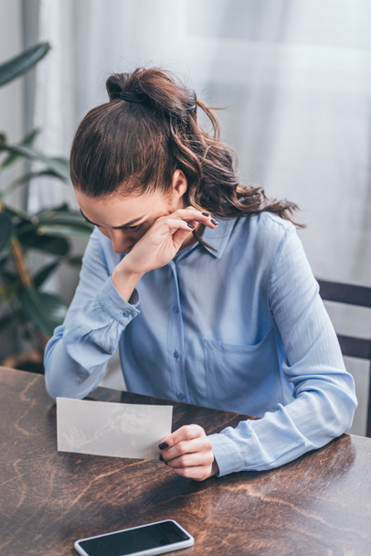 zdenerwowana Kobieta w niebieska bluzka siedzi przy stole z Fotografia, smartphone i płacz w domu żałoby koncepcja zaburzenia - Zdjęcie, obraz