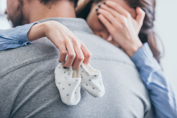 bijgesneden beeld van de vrouw, man knuffelen, holding van baby sokken en huilen op kamer, rouwende stoornis concept - Foto, afbeelding