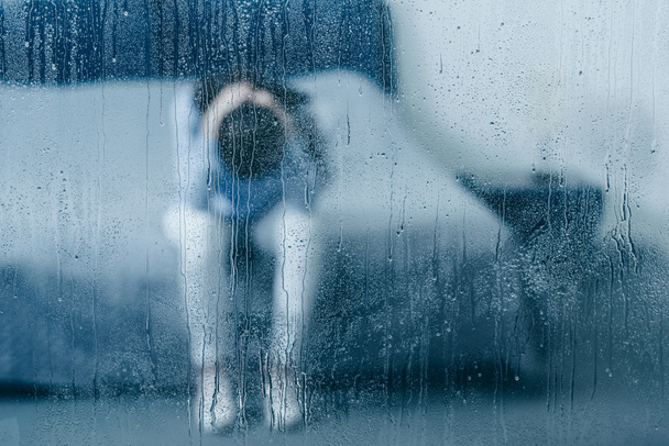 mujer deprimida sentada en la cama y sosteniendo la cabeza en las manos a través de la ventana con gotas de lluvia
 - Foto, Imagen