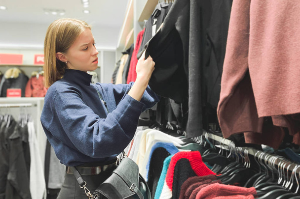 Aantrekkelijk meisje in een hippe kledingwinkel kiest kleding. Meisje kijkt naar wat kleren, winkelen in de boutique. Vrouw kijkt kleding in de winkel fashion. - Foto, afbeelding