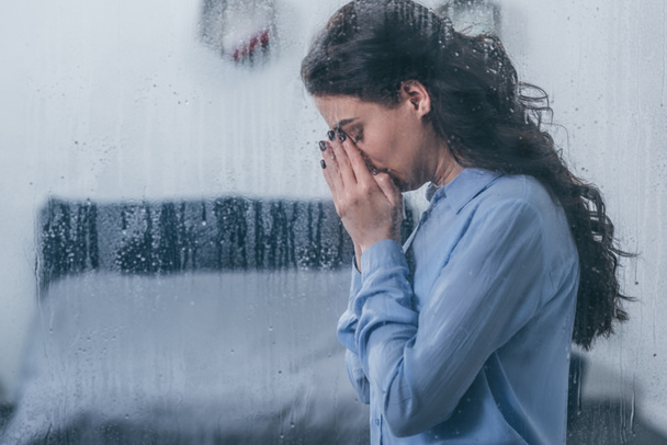 mujer deprimida cubriendo la cara con las manos y llorando en casa a través de la ventana con gotas de lluvia y espacio para copiar
 - Foto, Imagen