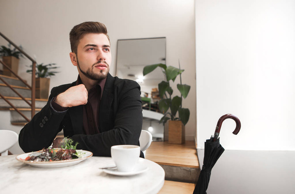 Bonito homem de negócios com barba e um terno sentado à mesa com uma salada e uma xícara de café e olhando pela janela. Retrato de um homem num fato de negócios num restaurante
. - Foto, Imagem