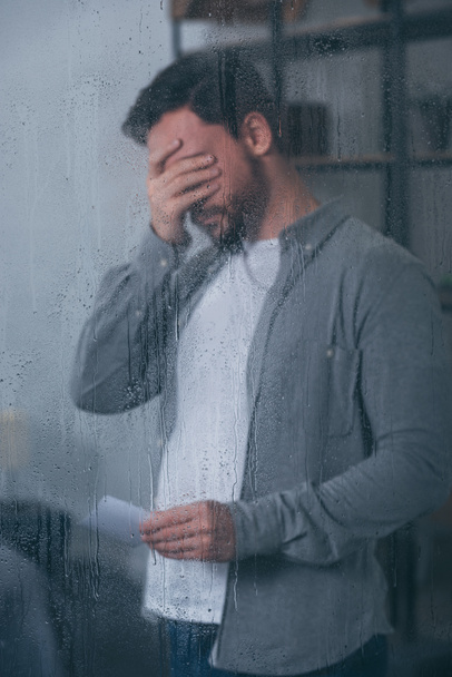 przygnębiony człowiek obejmujący twarz ręką, trzymając zdjęcie i płacz przez okno z deszczu - Zdjęcie, obraz