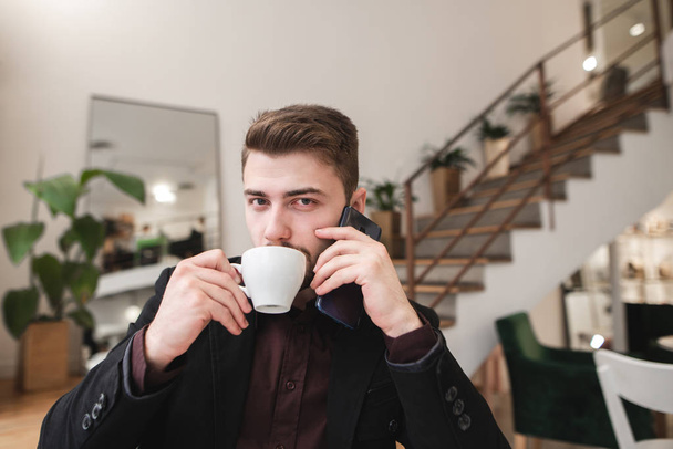 Handsome man in a suit drinks coffee in a cozy restaurant, speaks by phone and looks at the camera. Portrait of a business man talking on the phone in a cozy cafe. - Photo, Image