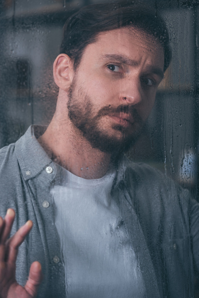 Красивый расстроенный мужчина трогает окно каплями дождя и смотрит в сторону
 - Фото, изображение