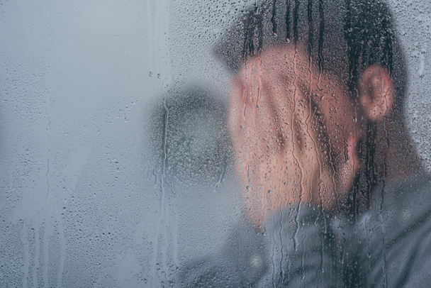 selektiver Fokus von Regentropfen auf Fenster mit Mann, der Gesicht bedeckt und im Hintergrund weint - Foto, Bild