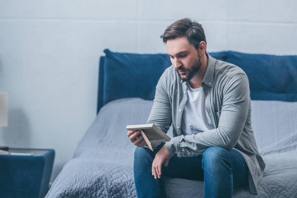 grieving man sitting on bed and looking at photo frame at home with copy space - Photo, Image
