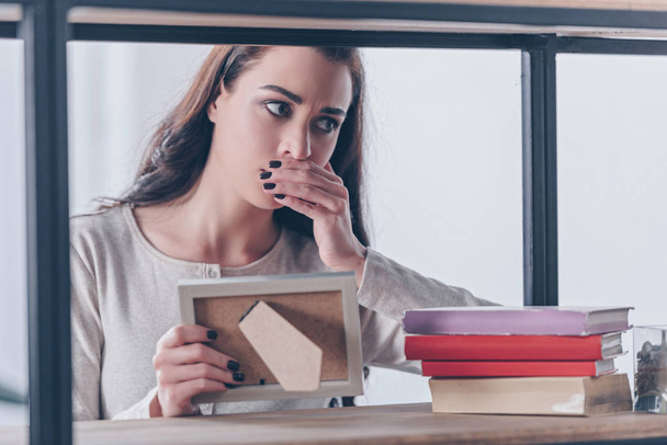 grieving woman covering mouth with hand while holding picture frame at home - Photo, Image