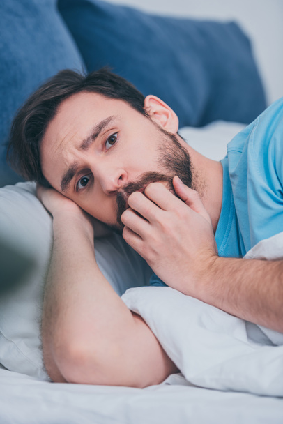 scared man lying in bed, looking at camera and covering mouth with hand - Photo, Image