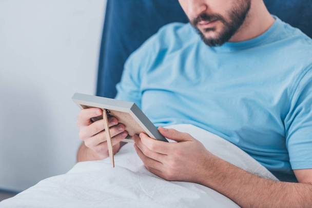 cropped view of bearded man looking at photo frame in bed - Fotó, kép