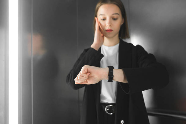 Young woman in the elevator is being late. Junior employee is looking at her watch understanding she is late. Businesswoman in a rush. - 写真・画像