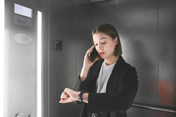 La joven empresaria ocupada está mirando su reloj y hablando por teléfono en el ascensor. Empleada atada está comprobando el tiempo de su reloj mientras habla por su teléfono
. - Foto, Imagen