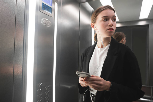 Businesswoman está echando un vistazo en el ascensor utilizando su teléfono inteligente y auriculares. Retrato de una trabajadora de oficina en el ascensor usando sus aparatos y mirando a la puerta
. - Foto, Imagen