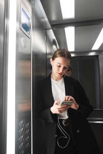 Вертикальна крупним планом фотографія бізнес-леді, що тримає смартфон у ліфті та слухає музику в навушниках. Портрет рук офісного працівника зі смартфоном та навушниками в ліфті
. - Фото, зображення