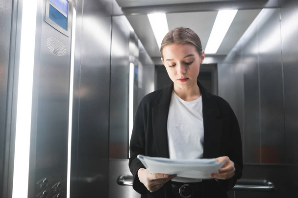Mladá podnikatelka je soustředěna při čtení dokumentů ve výtahu. Portrét pečlivé ženské úřednice kontrola a číst noviny ve výtahu. - Fotografie, Obrázek