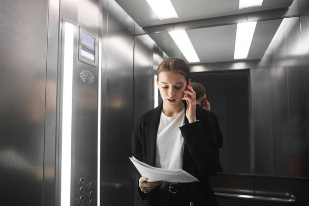 Расслабленная деловая женщина разговаривает по смартфону и читает документы в лифте. Концентрированная офисная работница проверяет газеты и разговаривает с боссом по телефону
. - Фото, изображение