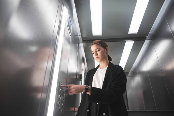 黒のスーツを身に着けている美しい若い実業家は、エレベーターを使用しています。野心的な女性事務員、エレベーターのボタンを押すこと。背景の水平方向写真. - 写真・画像