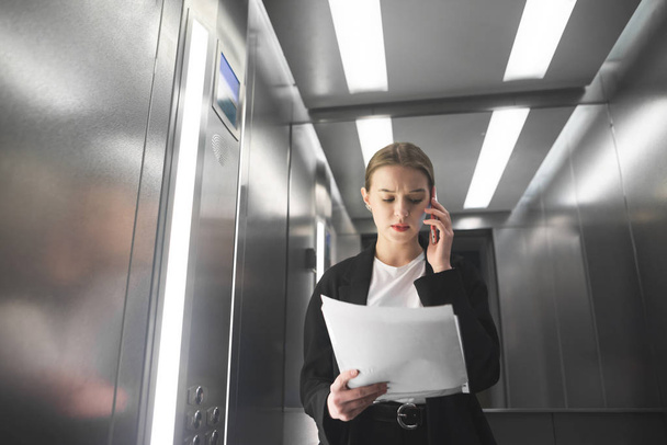 Serious businesswoman is talking by phone looking at papers in the elevator. Young female employee is reading the documents and talking by smartphone in the lift. - Foto, Imagen