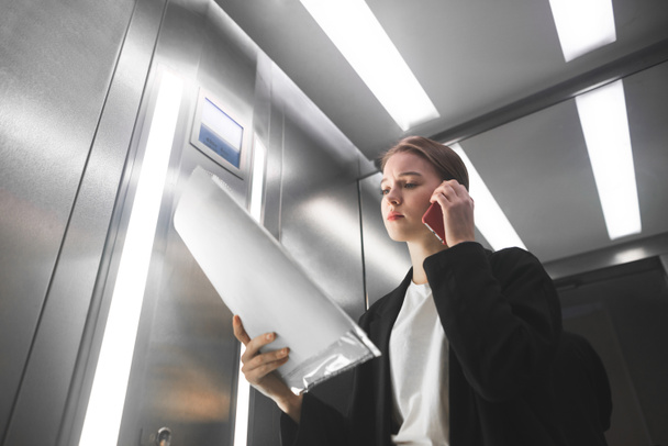 Concerned female office worker is speaking on the phone and checking the documents in the elevator. Young businesswoman is listening to the smartphone and reading the business papers in the lift. - Foto, imagen