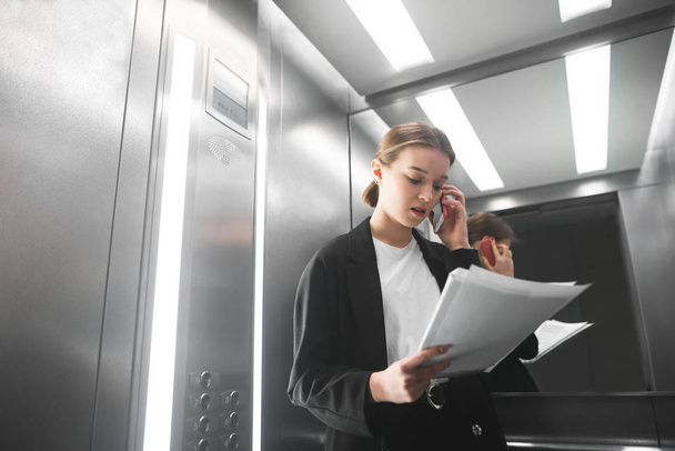 Busy businesswoman conference via smartphone reading the documents in the elevator. Concentrated office worker communicates with customer by phone and reads the papers. Background. - Photo, image