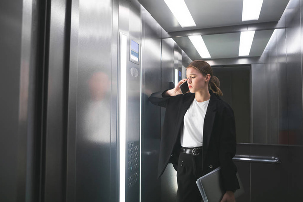 Die unzufriedene Geschäftsfrau spricht im Fahrstuhl auf ihrem Smartphone und hält den Laptop in der Hand. Frustrierte Büroangestellte hat Arbeitsprobleme und telefoniert mit ihrem Notizbuch. - Foto, Bild