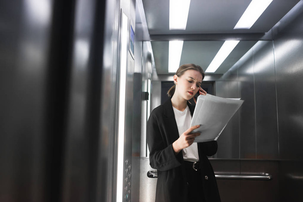 Serious female office worker talking on her smartphone and checking the documentation in the elevator. Young businesswoman reading the papers and communicate by phone. - Foto, afbeelding
