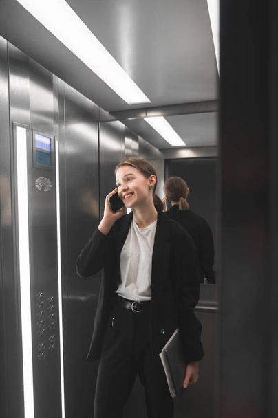 Joven mujer de negocios sonriente hablando por teléfono en ascensor sosteniendo portátil en la mano. Foto vertical de una trabajadora de oficina conversando por teléfono en ascensor con una amplia sonrisa. Satisfecho mirada feliz
. - Foto, Imagen