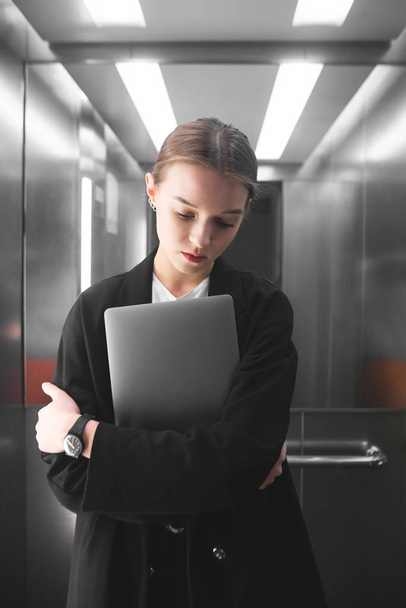 Молода бізнес-леді зосереджена і вдумлива, тримаючи ноутбук в ліфті. Портрет старанного працівника жіночого офісу, який дивиться вниз, думає і тримає свій записник в ліфті
. - Фото, зображення