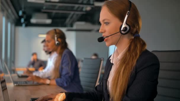 Call center employees working in deal office. Young handsome lady in headset on first plan talking with customer. Modern business room with beautiful view. Slow motion close-up. - Footage, Video