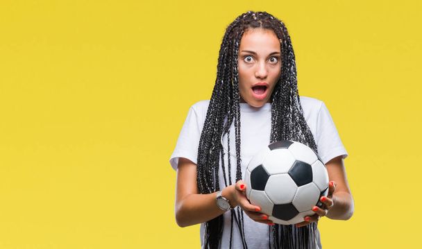 Молодая афроамериканка с плетеными волосами держит футбольный мяч на изолированном фоне, напуганная от удивления лицом, напуганная и взволнованная выражением страха
 - Фото, изображение