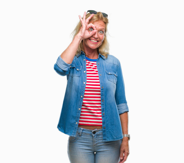 Μέσης ηλικίας ξανθιά γυναίκα απομονωμένη φόντο κάνει εντάξει χειρονομία με το χέρι, χαμογελώντας, το μάτι ψάχνει μέσα από τα δάχτυλα με χαρούμενο πρόσωπο. - Φωτογραφία, εικόνα