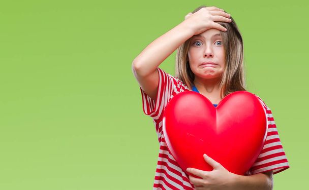 Young beautiful girl holding red heart over isolated background stressed with hand on head, shocked with shame and surprise face, angry and frustrated. Fear and upset for mistake. - Foto, imagen