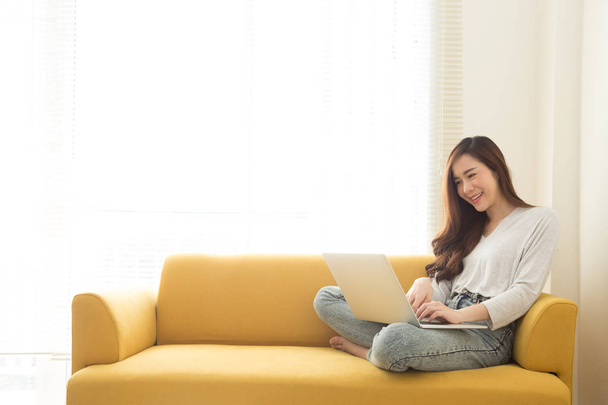 Young Asian woman sitting with laptop on yellow couch in home office. Concept of remote workplace and working at home - Photo, Image