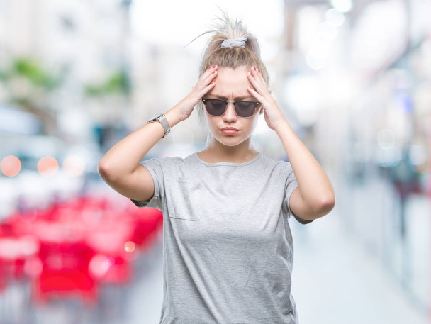 Young blonde woman wearing sunglasses over isolated background suffering from headache desperate and stressed because pain and migraine. Hands on head. - 写真・画像