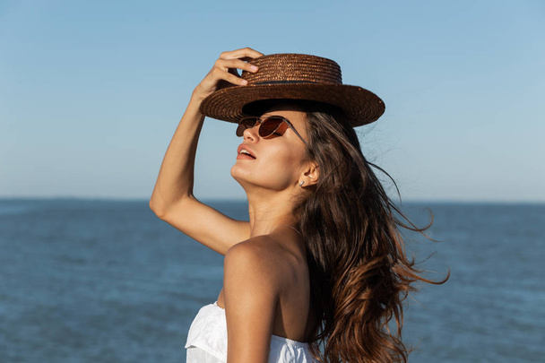Portrait of stunning dark-haired girl  in white dress, sunglasses and brown hat near the sea on a sunny day. - Foto, afbeelding