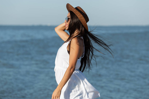 Portrait of stunning dark-haired girl  in white dress, sunglasses and brown hat near the sea on a sunny windy day. - Фото, изображение