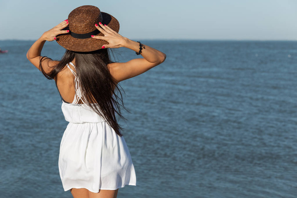 Gorgeous brunette girl with long hair in white dress and brown hat stand near the sea on a sunny day. - Photo, Image
