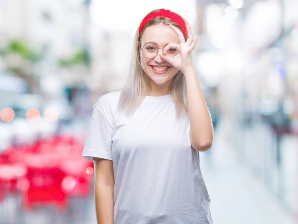 Giovane donna bionda indossa occhiali su sfondo isolato facendo gesto ok con la mano sorridente, occhio guardando attraverso le dita con il viso felice
. - Foto, immagini