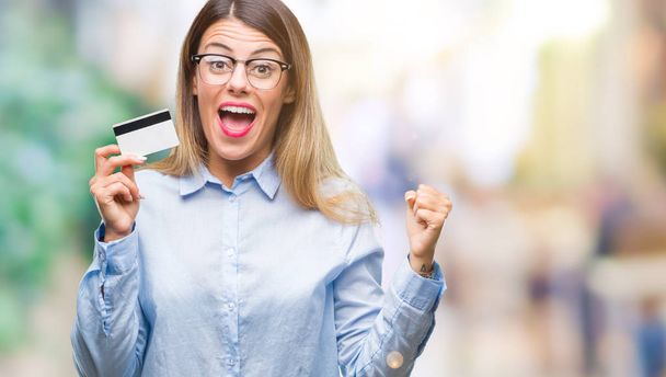 Young beautiful business woman holding credit card over isolated background screaming proud and celebrating victory and success very excited, cheering emotion - Photo, Image