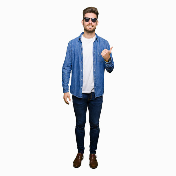 Mladý pohledný elegantní muž na sobě džínové sako dělá šťastné palec nahoru gesto rukou. Schvalování výraz s ukazuje úspěch při pohledu na fotoaparát. - Fotografie, Obrázek