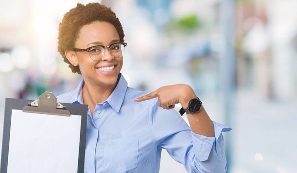 junge afrikanisch-amerikanische Geschäftsfrau hält Klemmbrett über isoliertem Hintergrund mit überraschendem Gesicht, das mit dem Finger auf sich selbst zeigt - Foto, Bild