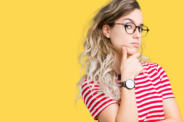 Beautiful young blonde woman wearing glasses over isolated background with hand on chin thinking about question, pensive expression. Smiling with thoughtful face. Doubt concept. - Photo, Image
