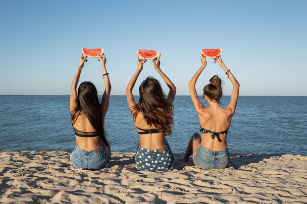 Back view of  three  dark-haired girls in jean shorts sitting on the sand near the sea holding watermelon slices over their heads - Foto, immagini