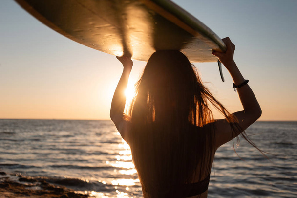 Dark-haired girl in a swimsuit holds a surfboard over her head on the sandy beach near the sea on the sunset - Фото, изображение