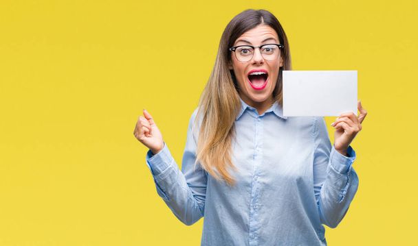 Young beautiful business woman holding blank card over isolated background screaming proud and celebrating victory and success very excited, cheering emotion - Photo, image