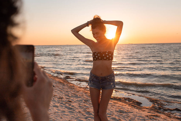 Girl make a photo of slim dark-haired girl in a swimsuit and shorts on the sandy beach near the sea on the sunset - Foto, Imagem
